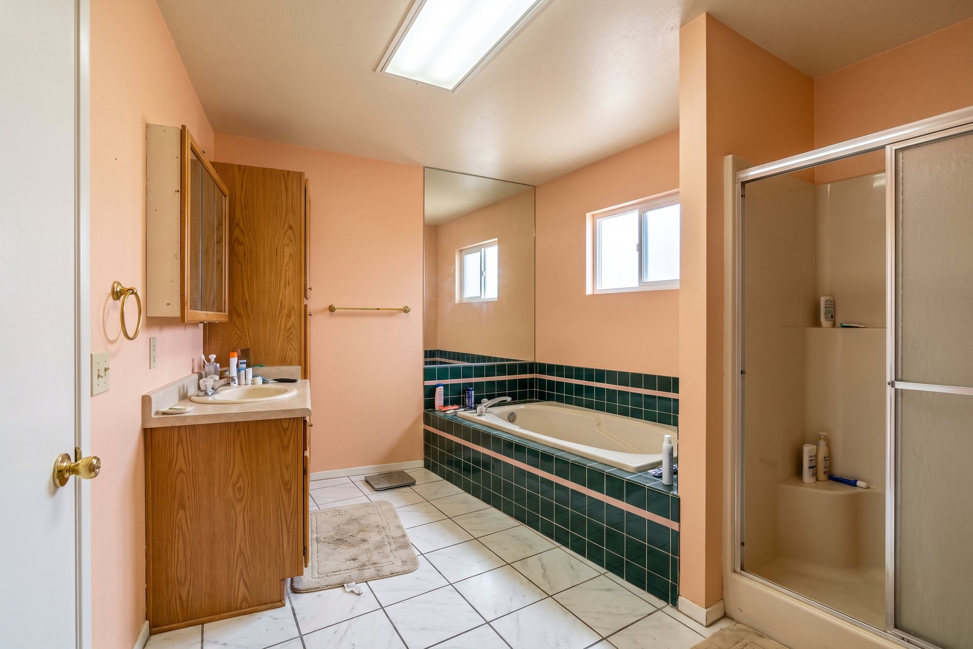 Before: a 1980s primary bathroom with a green-tiled platform tub under small frosted windows, a mirrored wall, a short oak vanity and a cramped fiberglass shower