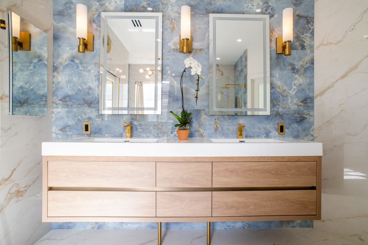 Double vanity with blue tile accent wall, two backlit mirrors, brass sconces, and matching brass faucets