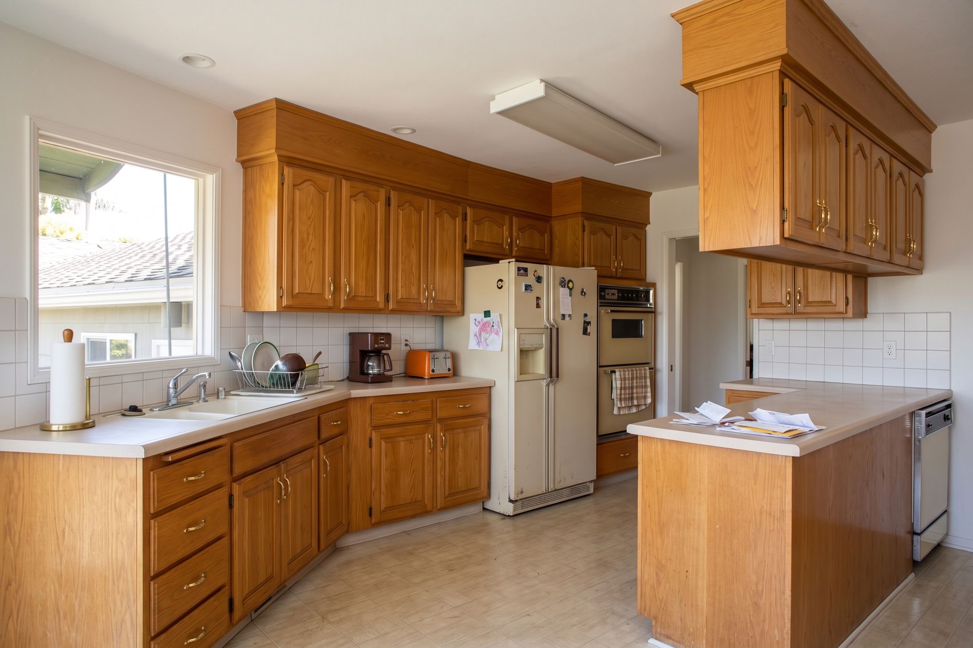 Before: the same Point Loma kitchen closed off by a peninsula with soffit cabinets above it, honey oak cabinets, laminate counters, plain white ceramic tile, an almond refrigerator wedged beside the oven and a fluorescent box light
