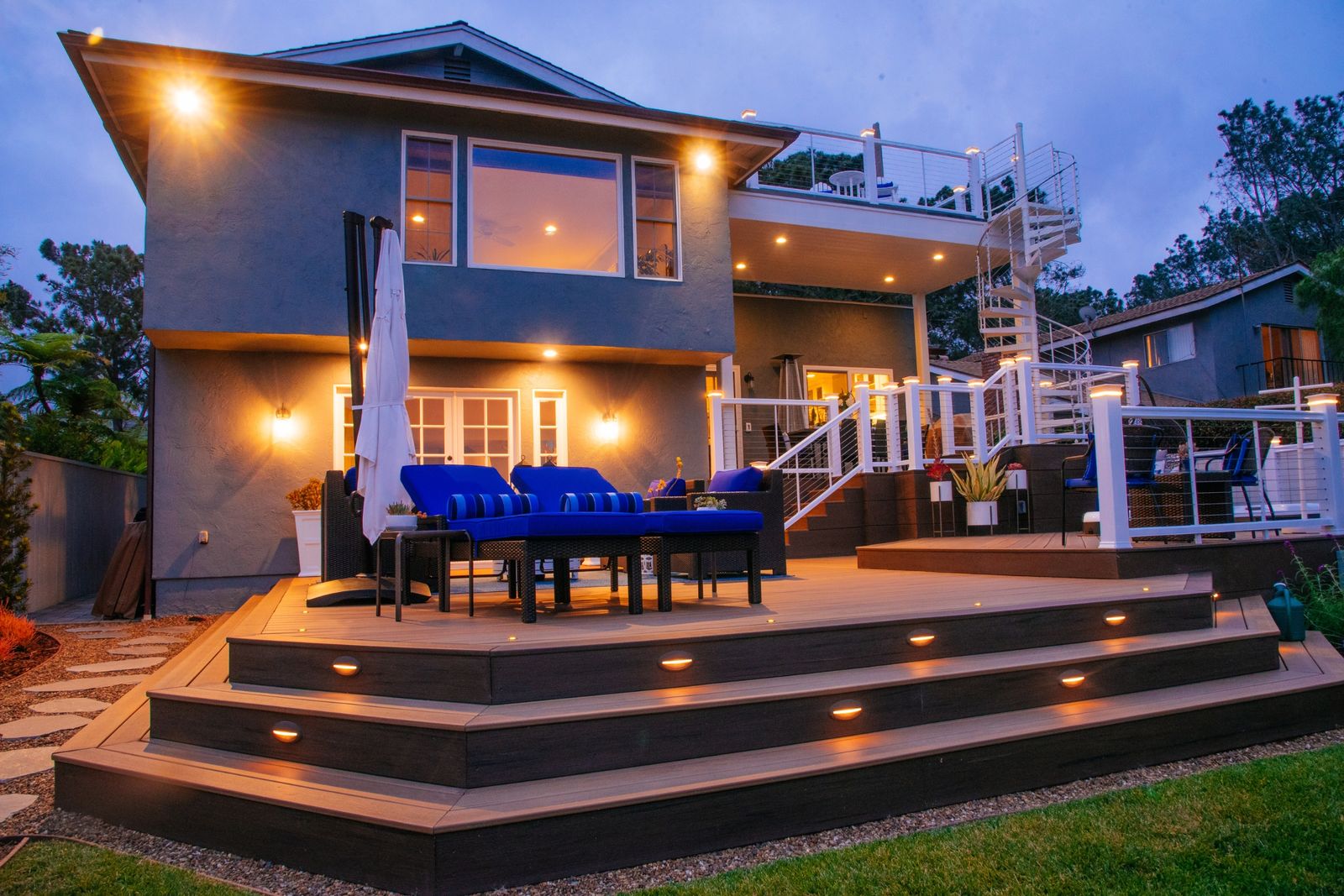 Point Loma three-level wood deck at dusk, lit steps and blue seating beside a two-story home