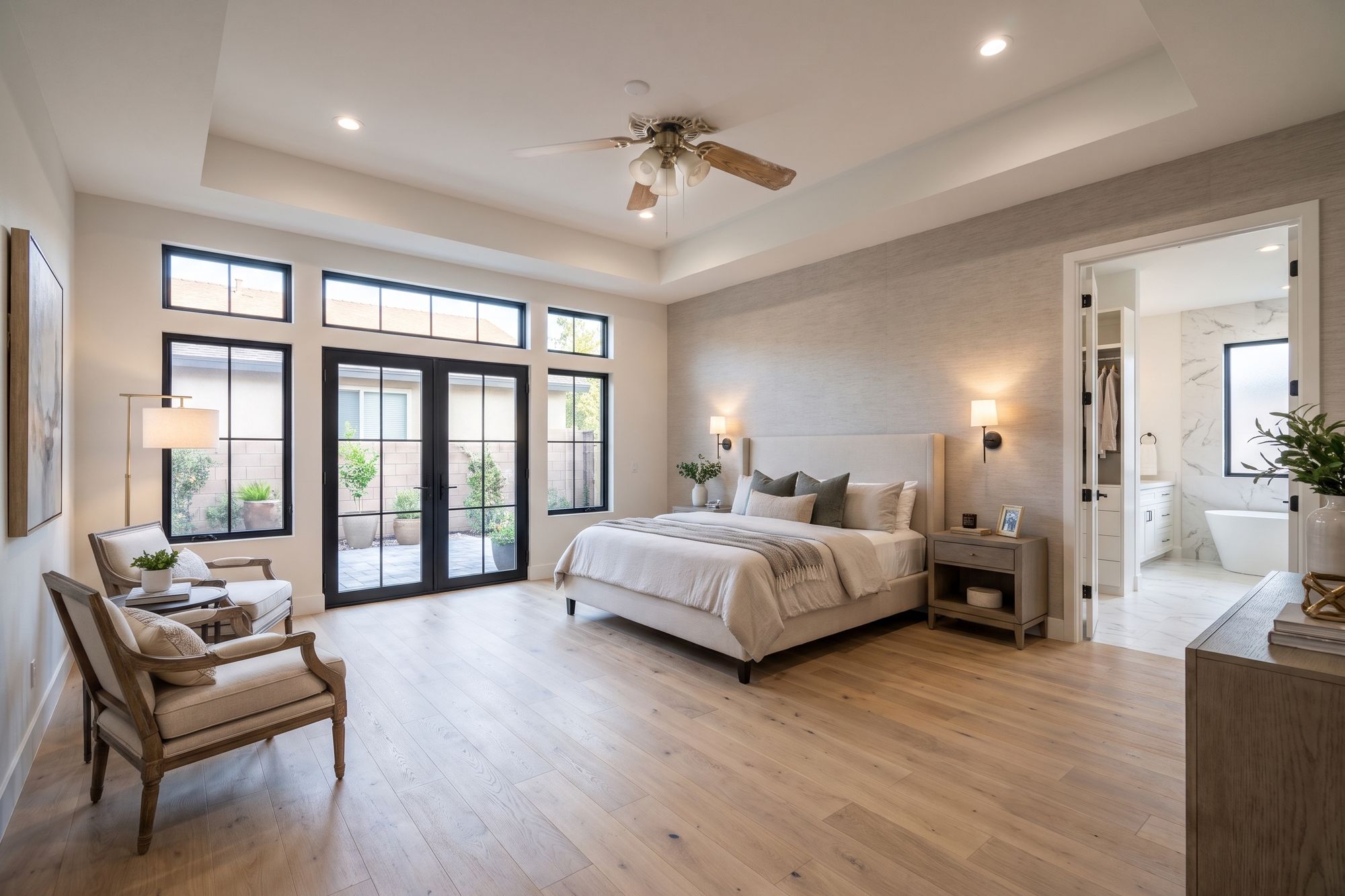 After: the same bedroom extended into a primary suite with a tray ceiling, tall black-framed windows and French doors to a patio, a sitting area, white oak floors and an open doorway to the walk-in closet and marble bath