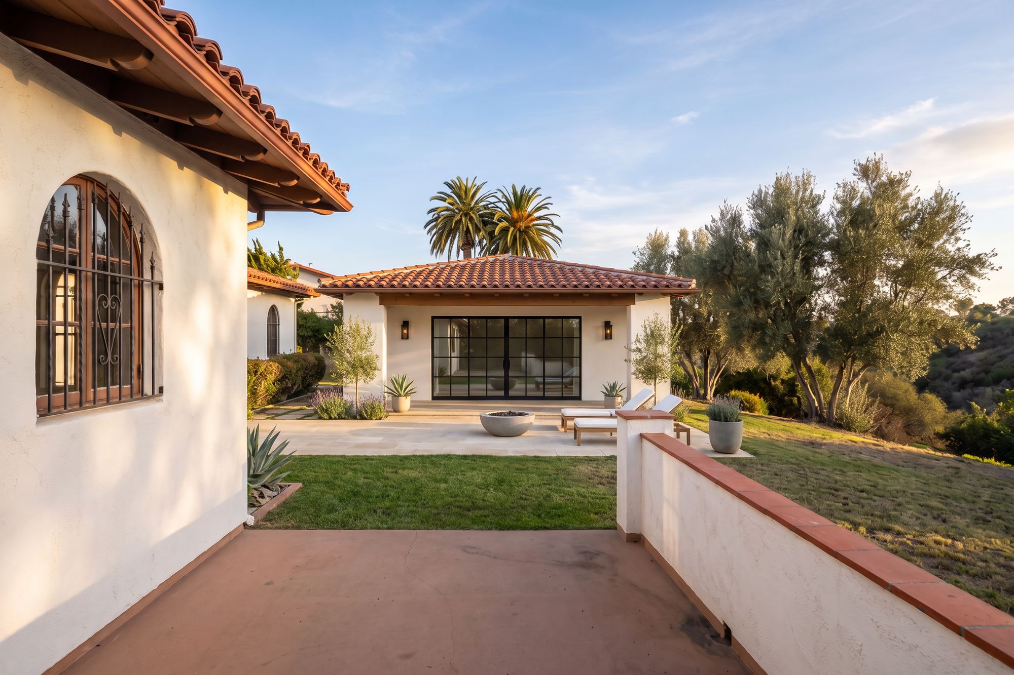 After: the same backyard with a custom modern Spanish ADU, white stucco and clay tile, a deep loggia with wood beams and a black steel glass wall, a limestone terrace with a fire bowl and loungers, olive trees and a fresh lawn