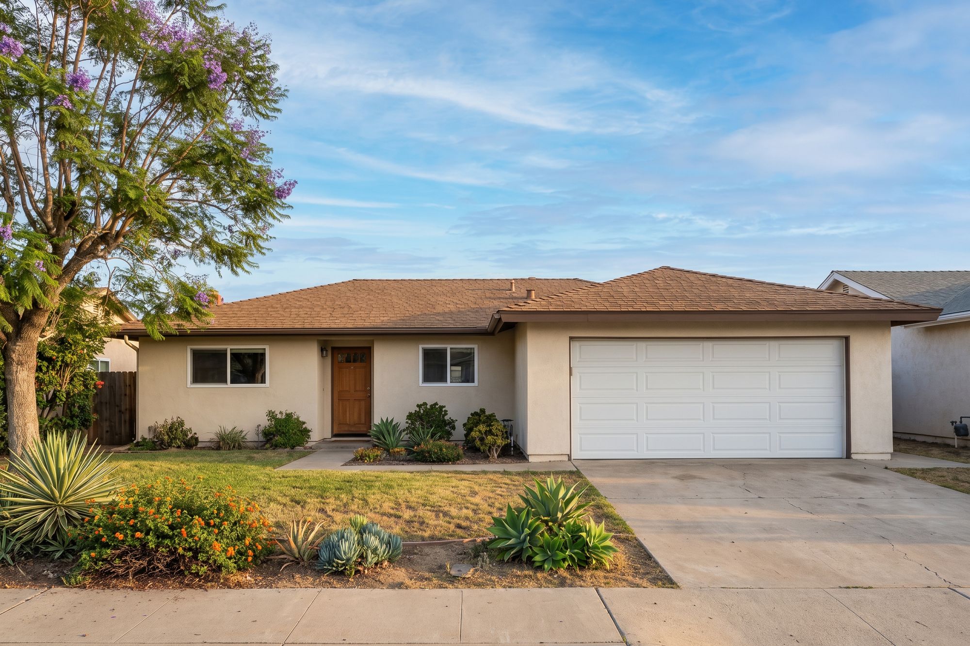 Before: a single-story 1970s San Diego ranch home with beige stucco, a brown shingle roof, a white two-car garage door and a jacaranda tree