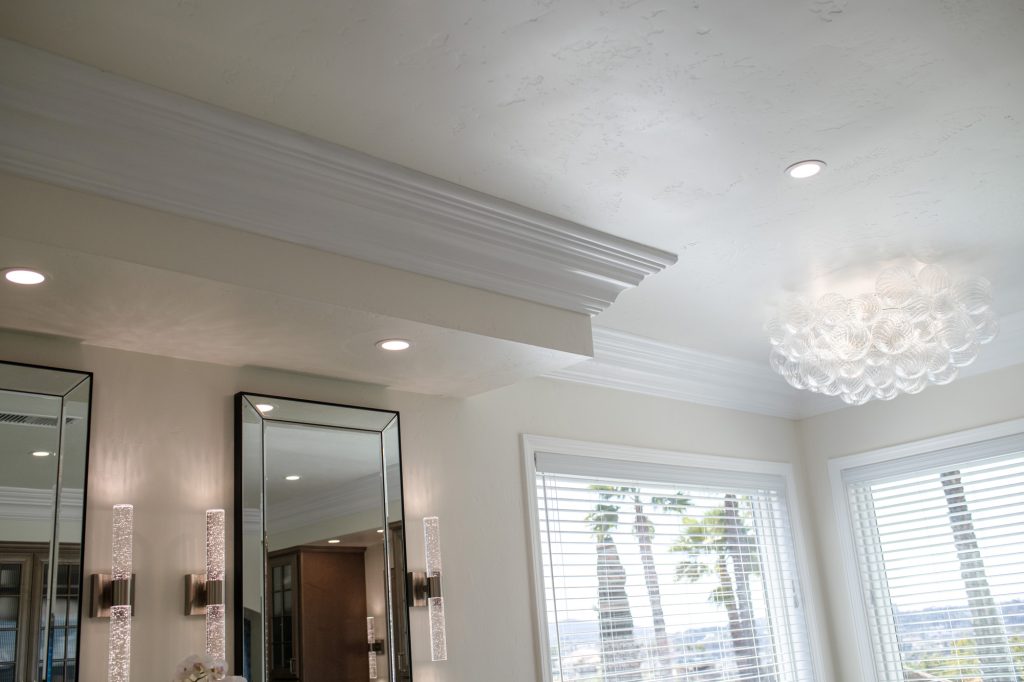 Crown-molded ceiling with glass-bubble chandelier, recessed lights, and palm-framed windows over the vanity area.