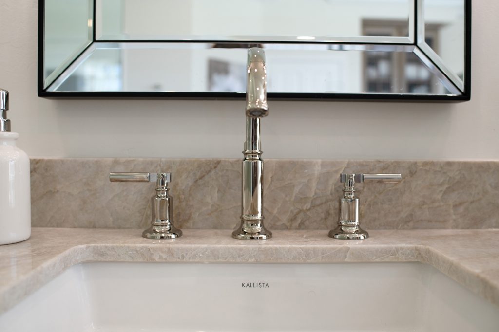 Close-up of polished-nickel Kallista widespread faucet set against a beveled mirror and quartzite backsplash.