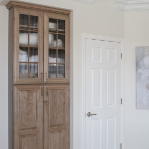 Maple-almond linen cabinet with ribbed-glass doors beside a white interior door and layered crown molding.