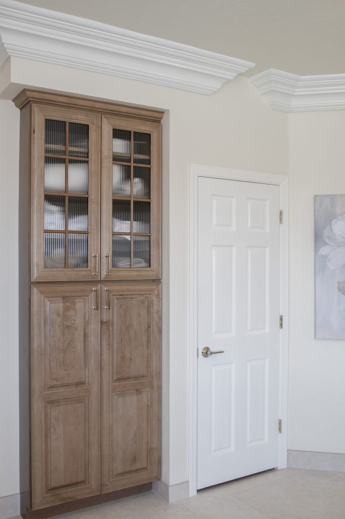 Maple-almond linen cabinet with ribbed-glass doors beside a white interior door and layered crown molding.