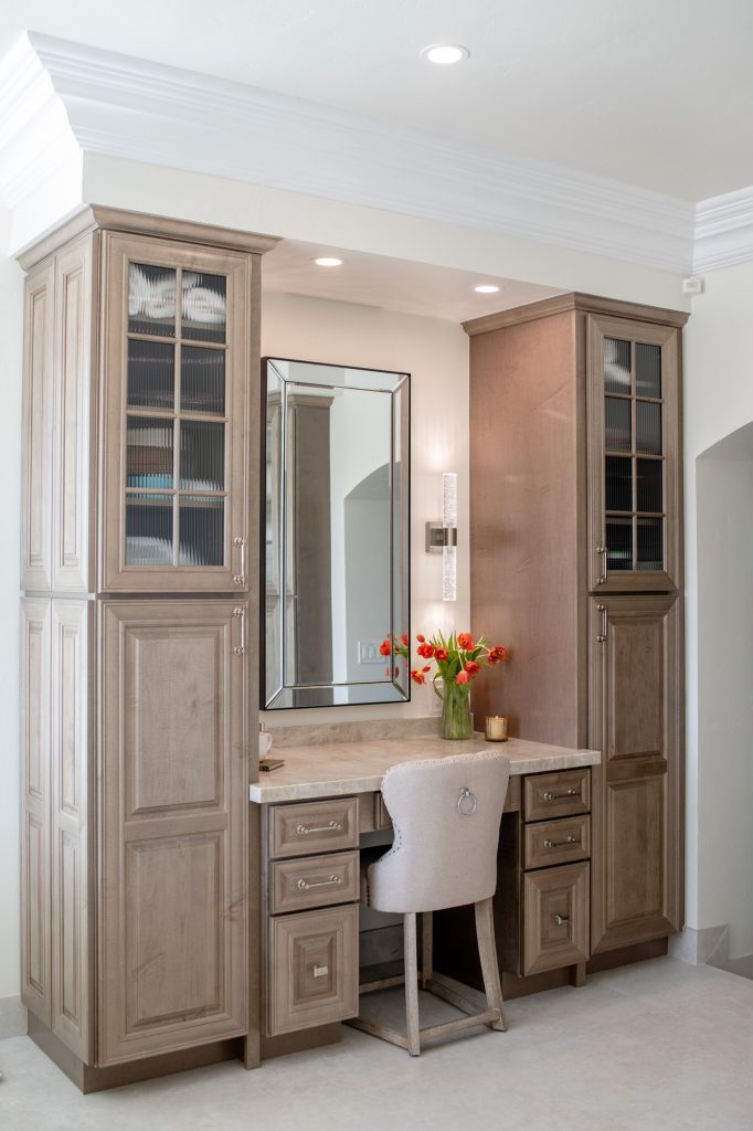 Full-height makeup vanity flanked by ribbed-glass tower cabinets, tulip bouquet and ring-pull chair beneath recessed lights.