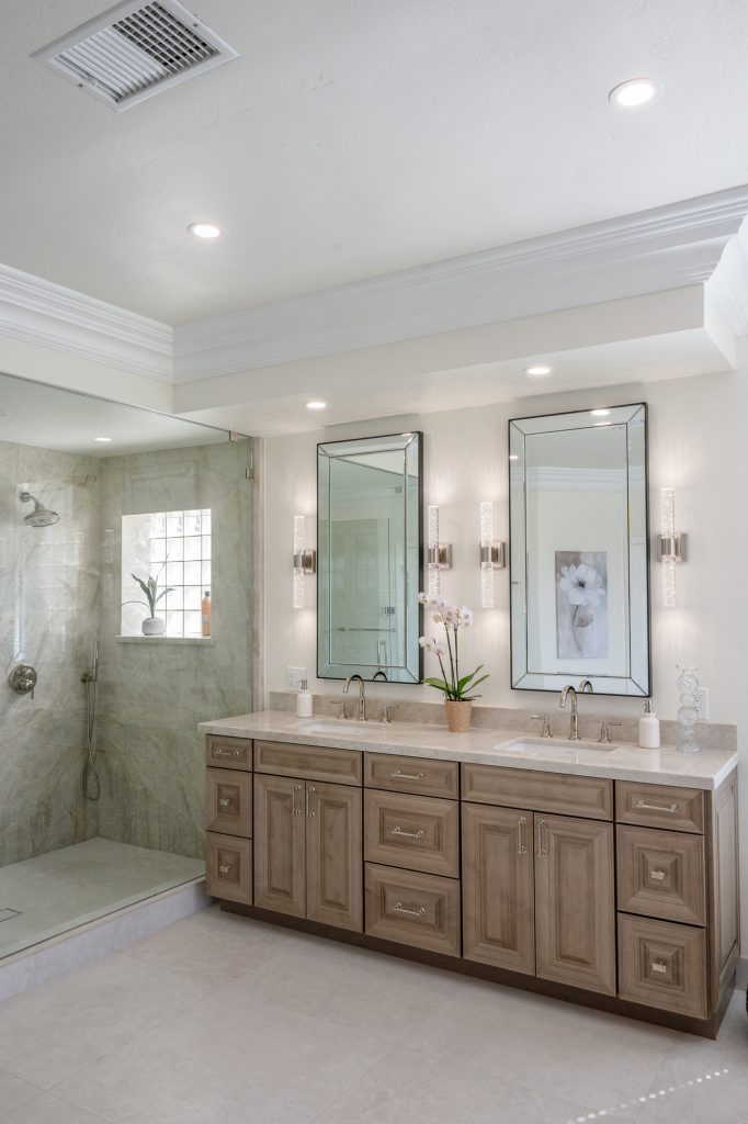 Wide view of double vanity with beveled mirrors and adjacent glass shower in the neutral luxury bath.