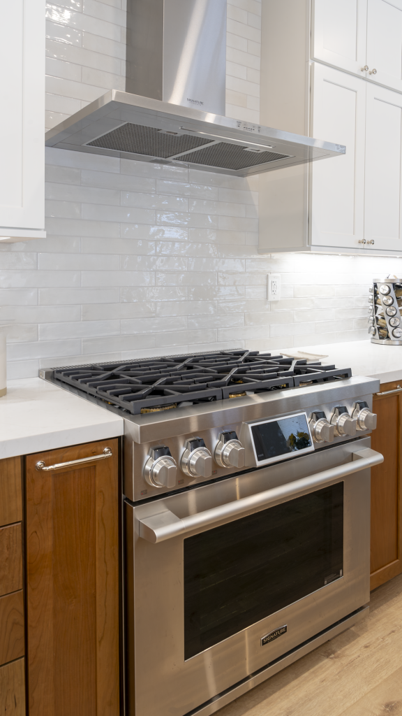Close-up of stainless professional gas range and hood framed by glossy ceramic backsplash and walnut cabinetry
