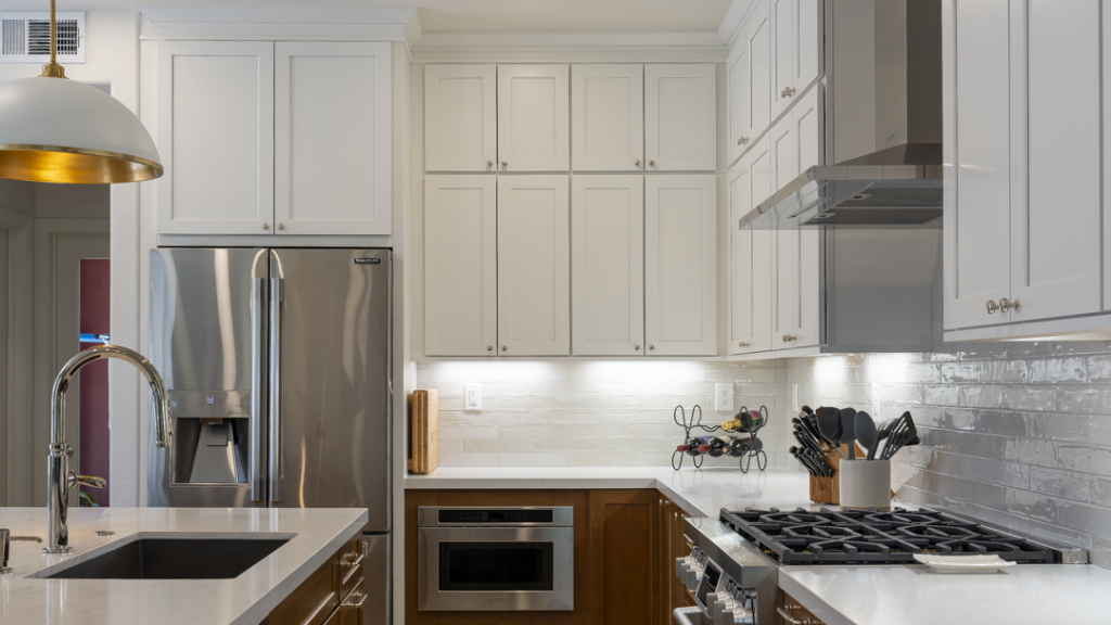 Close corner shot of fridge, microwave drawer, quartz counters, and two-tone cabinetry under under-cabinet lighting