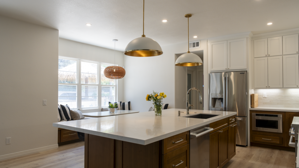Island sink view toward breakfast nook; quartz surfaces, brass pendant lights, stainless fridge, and light plank flooring