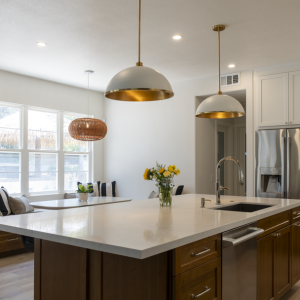 Island sink view toward breakfast nook; quartz surfaces, brass pendant lights, stainless fridge, and light plank flooring