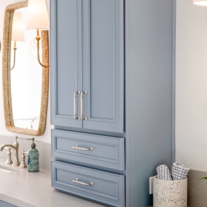 Blue linen tower cabinet with drawers on quartz countertop beside vanity mirror and lighting