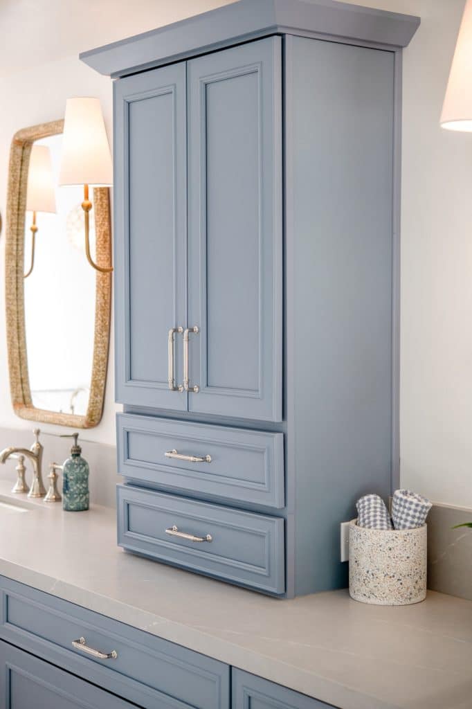 Blue linen tower cabinet with drawers on quartz countertop beside vanity mirror and lighting