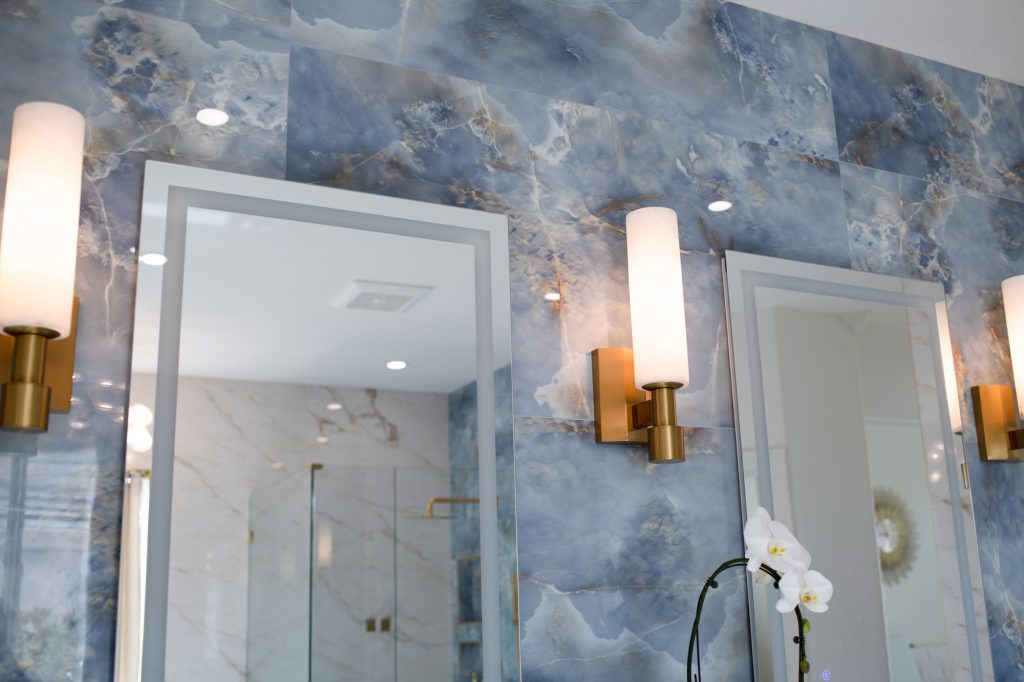 Brass wall sconces beside illuminated mirrors on a blue onyx-look tile feature wall