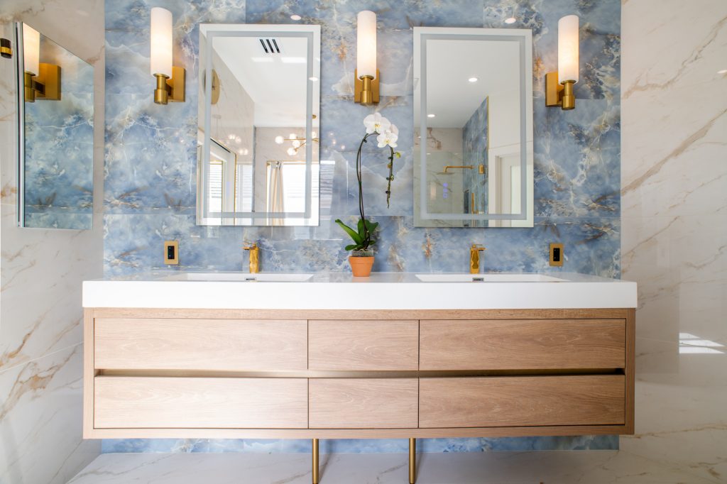 Double vanity with blue tile accent wall, two backlit mirrors, brass sconces, and matching brass faucets