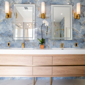 Double vanity with blue tile accent wall, two backlit mirrors, brass sconces, and matching brass faucets