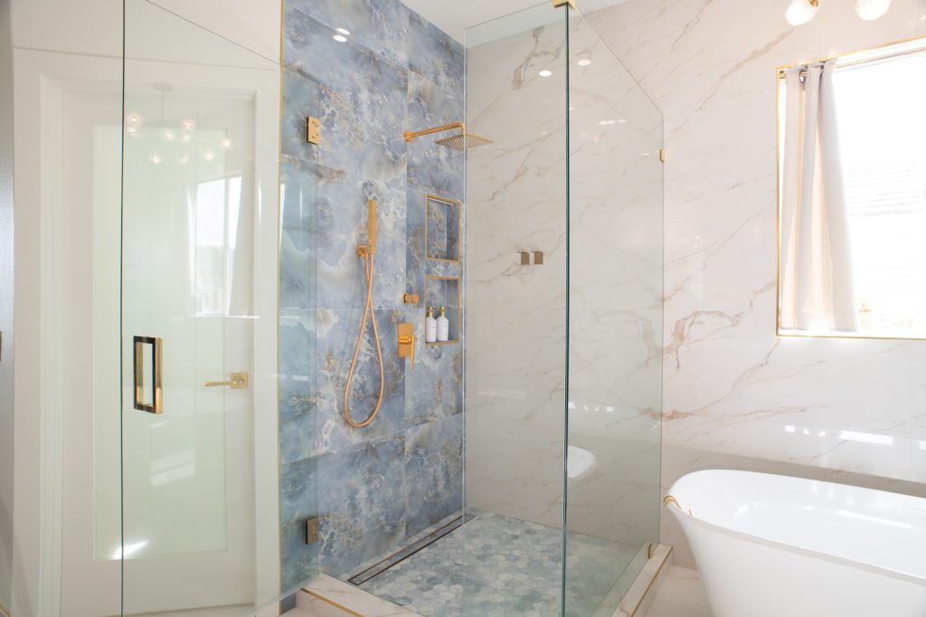 Glass-enclosed shower with a blue feature wall and brass fixtures next to a freestanding tub and marble-look tile walls