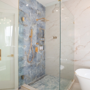 Glass-enclosed shower with a blue feature wall and brass fixtures next to a freestanding tub and marble-look tile walls