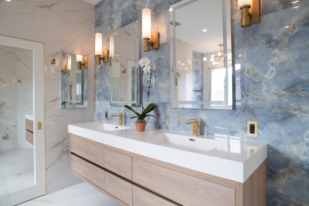 Modern double sink bathroom vanity with blue stone-look wall tile, brass sconces, and light wood cabinetry