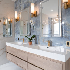 Modern double sink bathroom vanity with blue stone-look wall tile, brass sconces, and light wood cabinetry