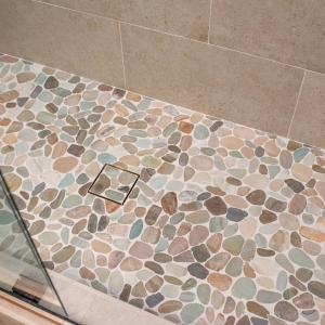 Sliced pebble shower floor with a square tile-in drain, seen past the glass door panel