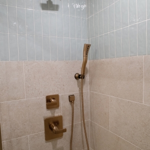 Square showerhead above a handheld wand, both in bronze, where the blue and beige tiles meet