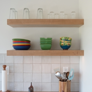 Two maple floating shelves holding glasses and colorful bowls above the backsplash